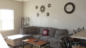 A living room with a gray sectional sofa, a coffee table, a dining table with chairs, a wall clock, and decorative mirrors.
