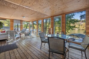En træterrasse med udendørs møbler, store vinduer og udsigt til et sø.