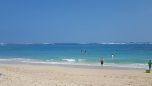 Ein Strand mit klarem, blauem Wasser, sandigem Ufer und Fußspuren, die ins Wasser führen.