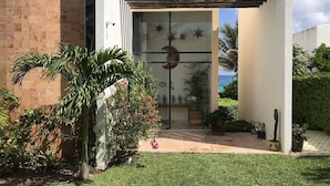 A modern building with a glass door, a small garden with palm trees, and a view of the sea. - Beautiful Oceanfront Mexican Villa with Infinity Pool, and Spectacular View (Isla Mujeres)
