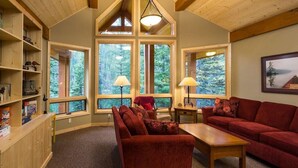 A cozy living room with a red sofa, a wooden coffee table, and a bookshelf. Large windows offer views of the forest.