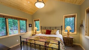A bedroom with a large bed, two bedside tables with lamps, and three windows offering a view of the forest.