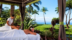 Un espace spa comprenant deux tables de massage, une personne s’occupant d’un client, et une vue sur l’océan et les palmiers.