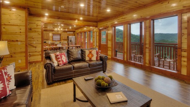 A wooden cabin interior with a leather sofa, a dining table, and a view of the outdoors. - A Heavenly View Too ~ 6 BR w/ Indoor Pool ~ Sleeps 28 ~ Incredible Views! (Sevierville)