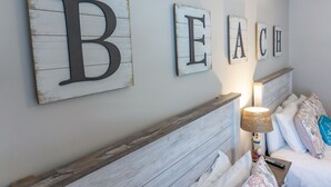 Quarto com cama, mesa de cabeceira e parede com letras de madeira formando a palavra 'BEACH'.