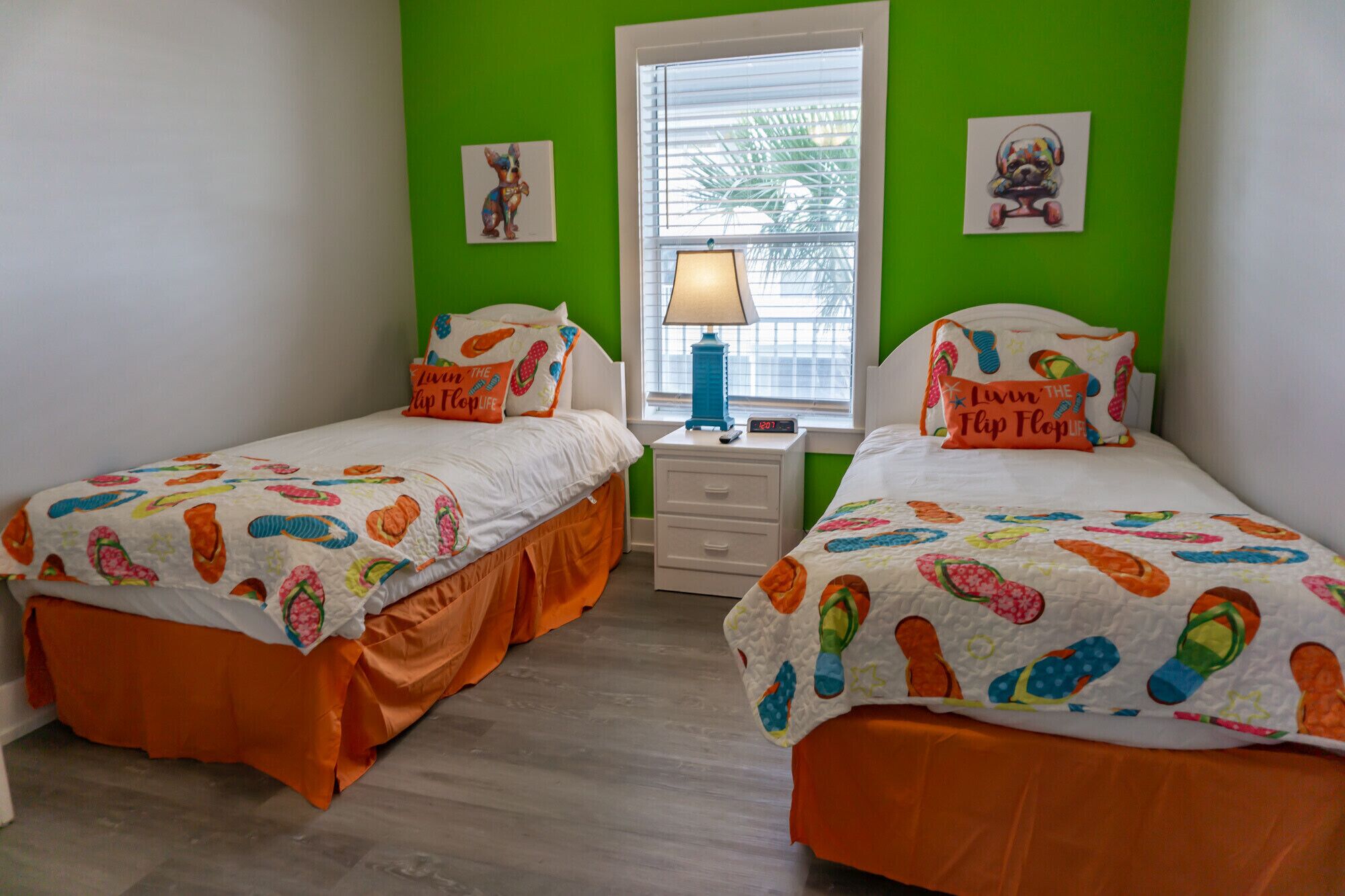 A room with two beds, each with a colorful bedspread featuring flip flops. There is a window with blinds, a lamp on a bedside table, and a clock.