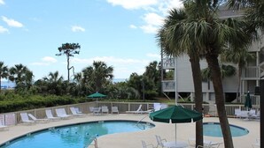A pool area with lounge chairs and umbrellas, surrounded by palm trees and a building with balconies. - Oceanfront condo, steps to beach & pool, across from Coligny Plaza! (Hilton Head)
