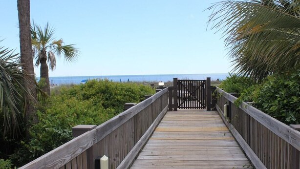 A wooden boardwalk leading to a gate, surrounded by greenery and palm trees, with a view of the sea in the distance. - Oceanfront condo, steps to beach & pool, across from Coligny Plaza! (Hilton Head)