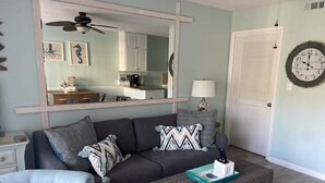A living room with a sofa, armchair, and ottoman, a large mirror, a clock, and a kitchen visible through the mirror. - Oceanfront condo, steps to beach & pool, across from Coligny Plaza! (Hilton Head)