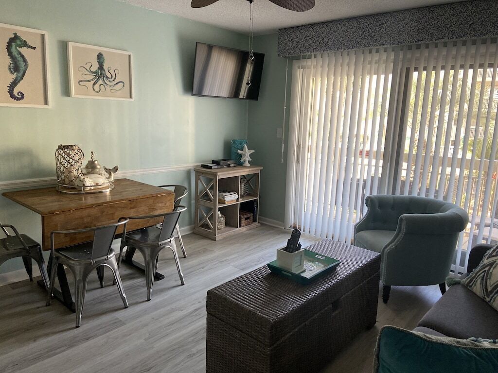 Un salon comprenant une table à manger en bois, des chaises en métal, une table basse en osier, un fauteuil vert et un téléviseur fixé au mur.