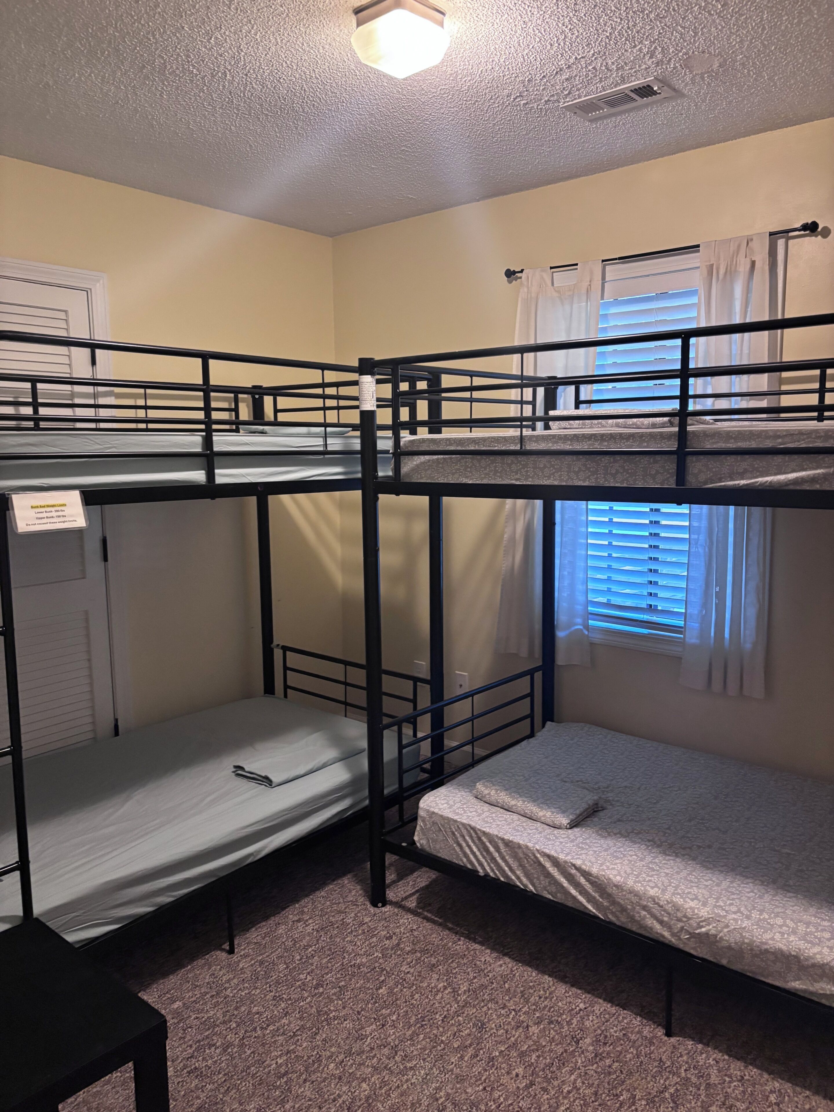 Une chambre avec trois lits superposés, une fenêtre avec des stores et un luminaire au plafond.