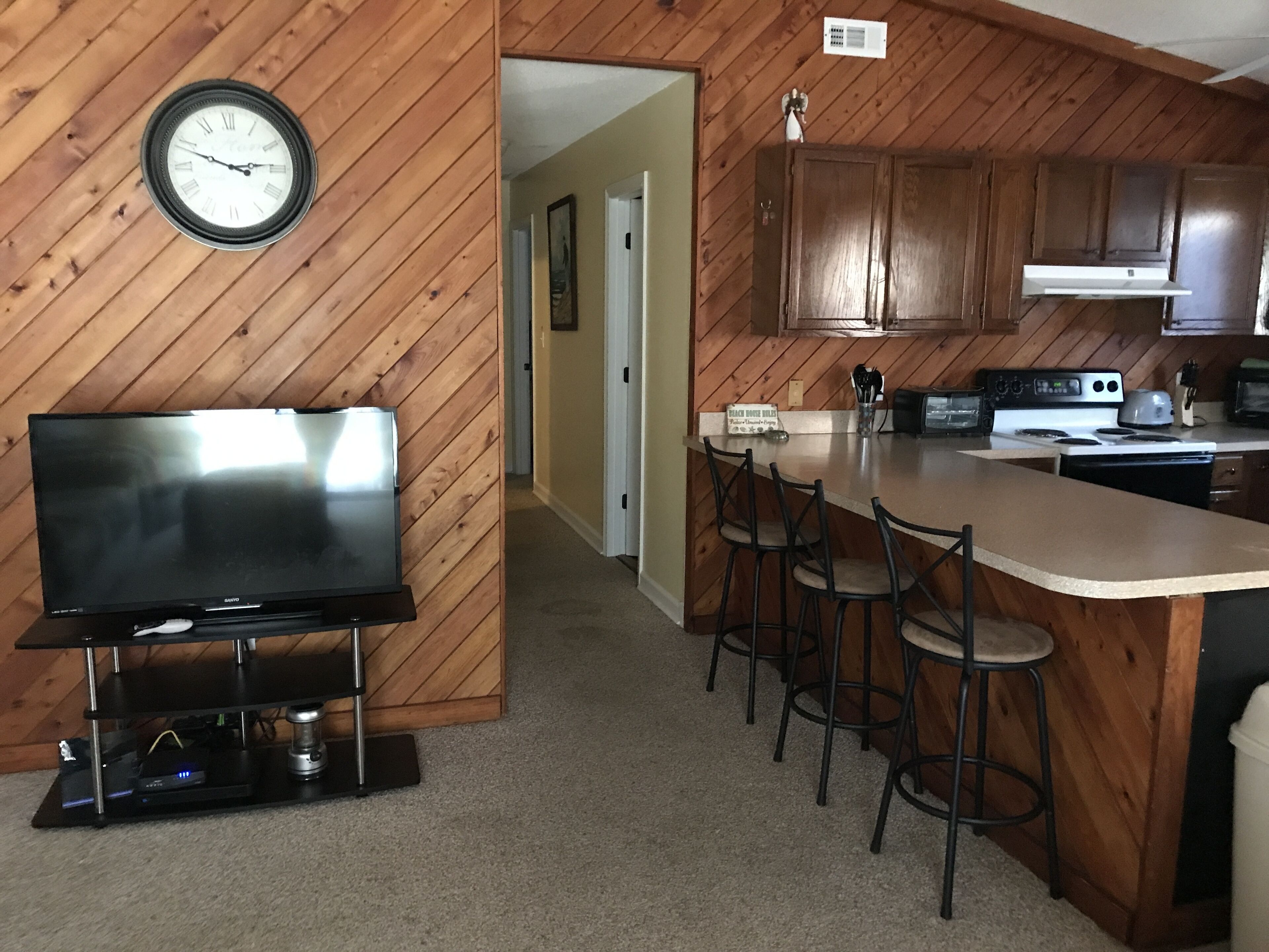 A kitchen with wooden cabinets, a built-in bar with stools, a clock on the wall, and a flat-screen TV.