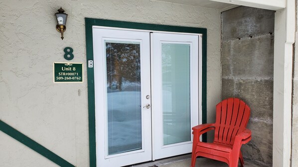 A double door with frosted glass panels, a green-framed door, and a red plastic chair outside. A green sign with "Unit 8" and a phone number is visible on the wall.