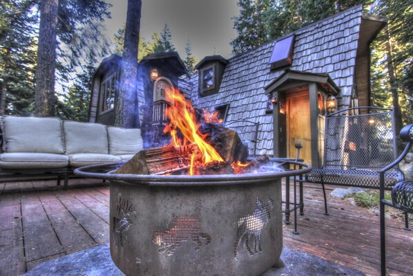 A fire pit with burning logs in a rustic outdoor setting, surrounded by wooden deck furniture and a stone building with a chimney.
