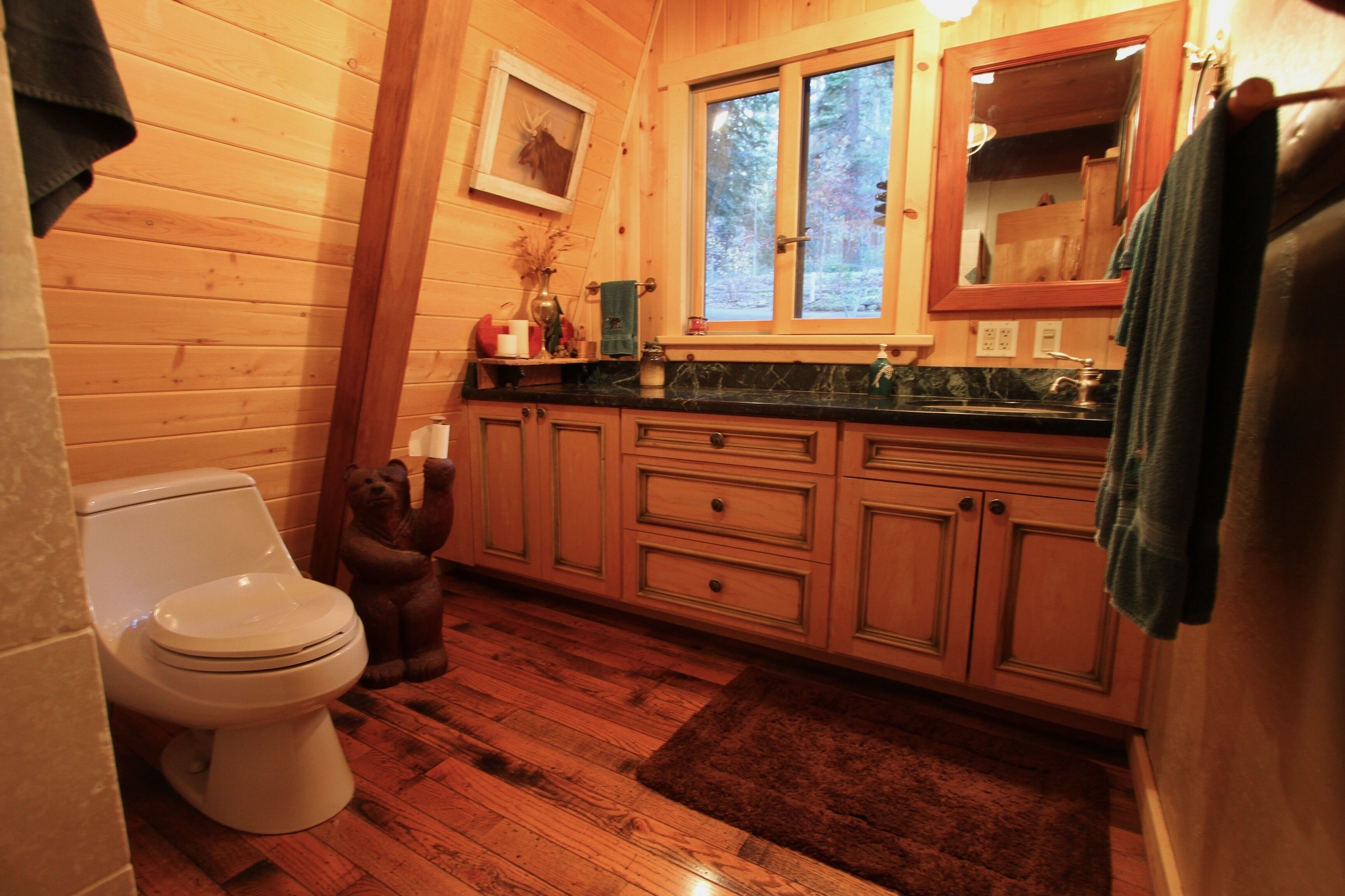 Ein Badezimmer einer Holzhütte mit Toilette, Holzschränken, einem Waschbecken und einem Fenster mit Blick auf Bäume.