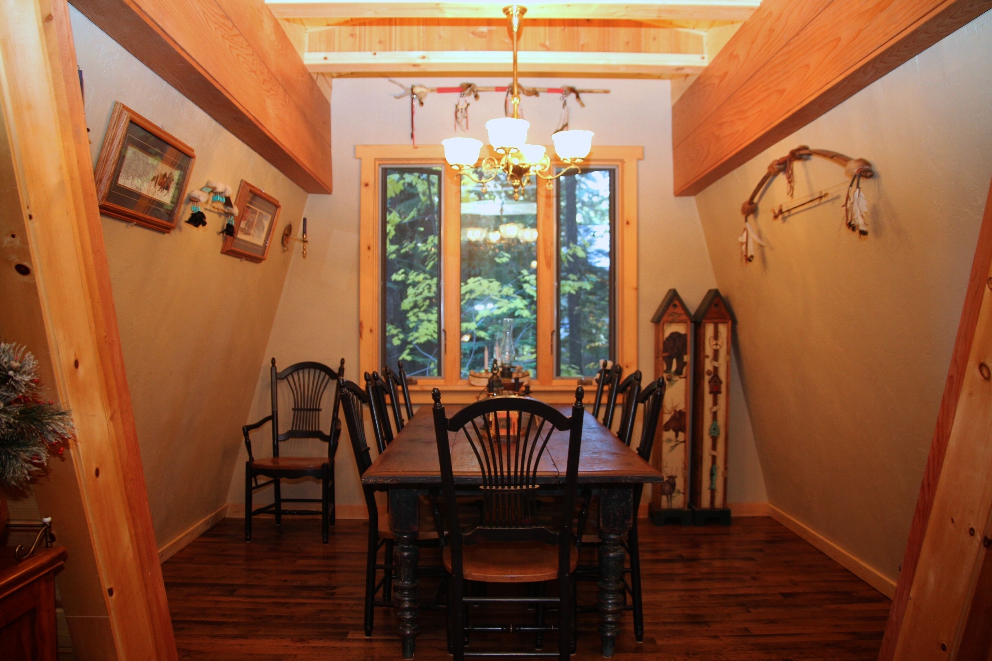 Une salle à manger avec une table et des chaises en bois, des tableaux encadrés au mur et une grande fenêtre donnant sur la forêt.
