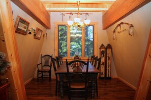 A dining room with a wooden table and chairs, framed pictures on the wall, and a large window with a view of trees.