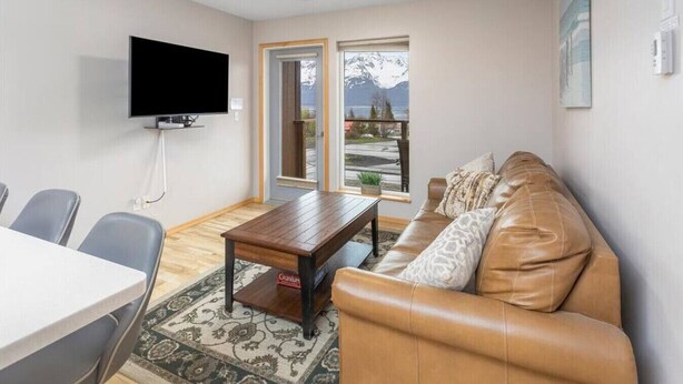 Una sala de estar con un sofá de cuero marrón, una mesa de centro de madera, un televisor de pantalla plana montado en la pared y una zona de comedor con una mesa blanca y sillas grises.