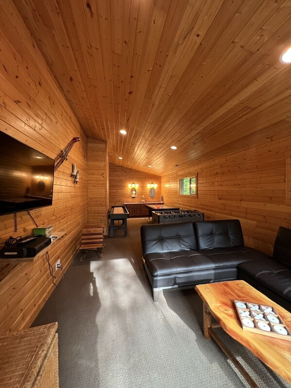 A cozy cabin interior with wooden walls, a black leather sofa, a wooden coffee table, and a foosball table in the background. - Theater, Hot Tub & River Access. This home was built for big groups! (Leavenworth)