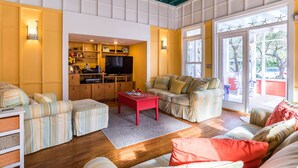 A cozy living room with a sofa set, a red coffee table, and a television. There is a kitchen area with a TV and various items on the counter. The room has a large window with a view of the outdoors. - 'Roger's Lighthouse' in the Heart of Seaside. Bikes included.  (seaside)