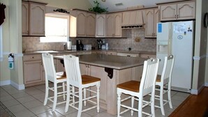 Een keuken met een groot eiland, witte houten stoelen, beige kastjes en een koelkast.