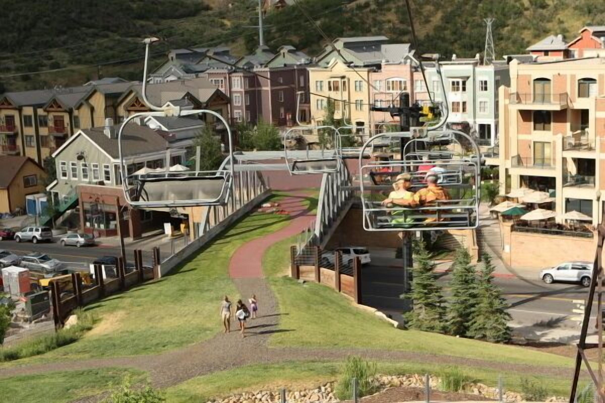 A ski lift with passengers, a pedestrian path, and residential buildings in the background.