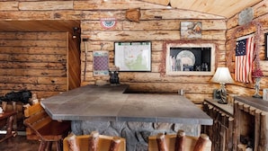 A rustic cabin interior with a large stone table, wooden chairs, a map on the wall, and an American flag.