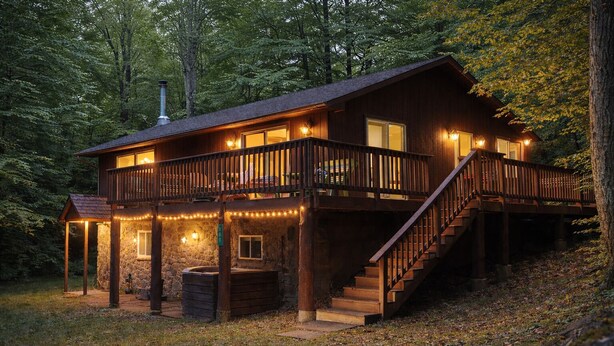 A two-story cabin with a balcony, stone foundation, and a wooden deck.