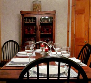 A dining table set with plates, glasses, and a candle. There are chairs around the table, and a wooden cabinet with glass doors in the background.