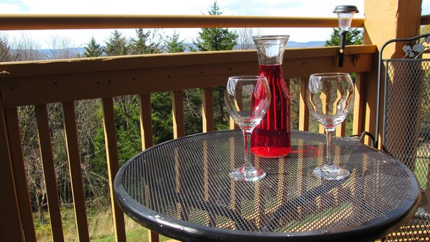 Een ronde metalen tafel met twee wijnglazen en een fles rode wijn op een balkon met houten leuningen en uitzicht op bomen en bergen.