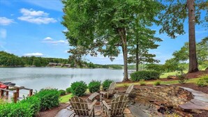A lakeside patio with Adirondack chairs, a stone fire pit, and a view of the lake and surrounding landscape.