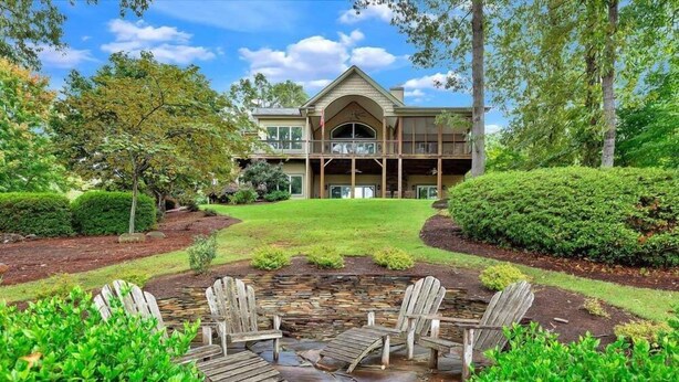 A large house with a balcony, surrounded by well-maintained gardens and a stone patio with Adirondack chairs.