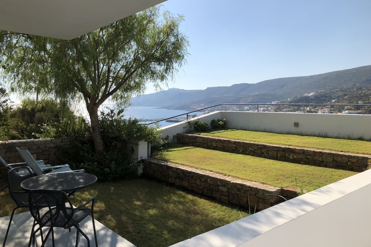 A balcony with a view of the sea and mountains, a stone wall, and a table with chairs.