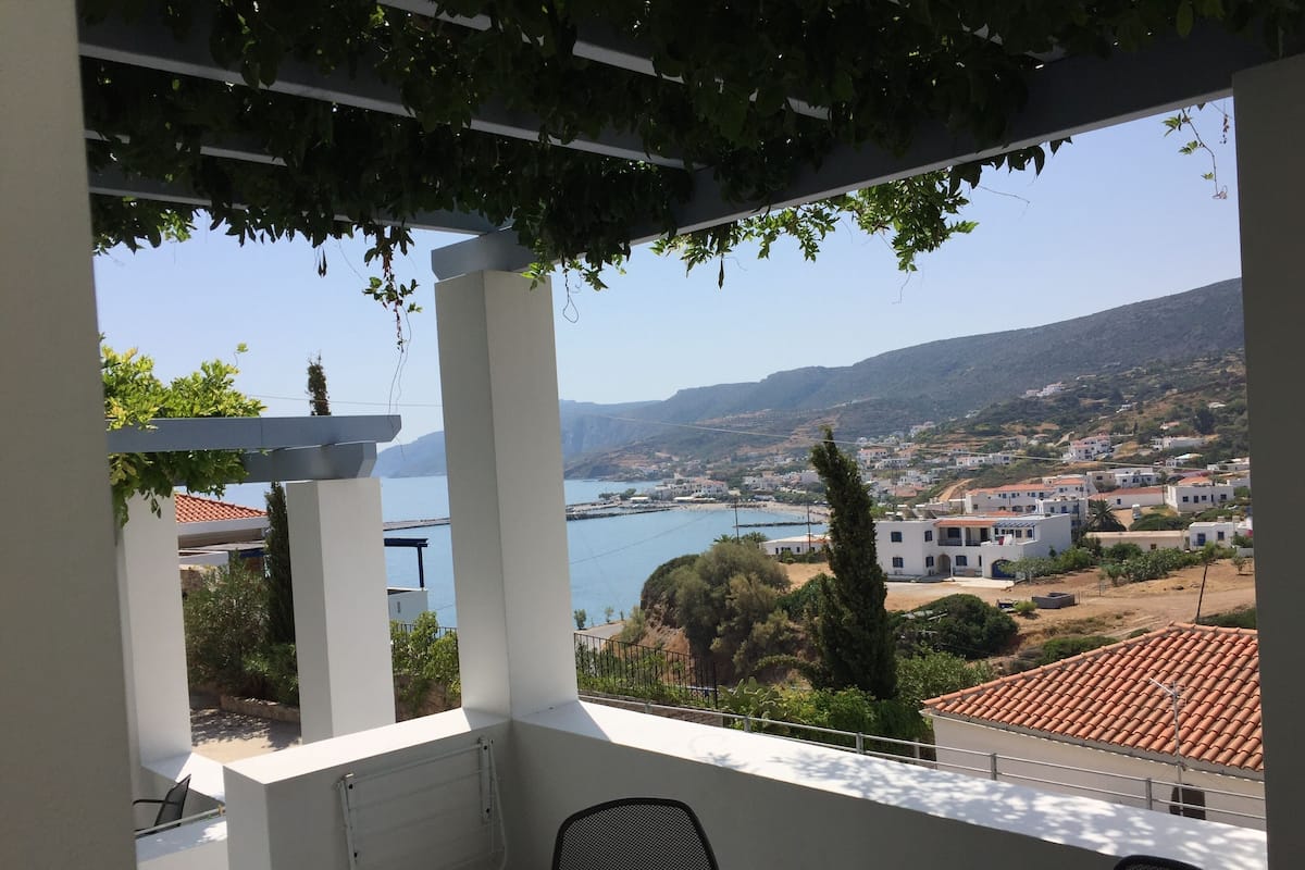 A balcony with a view of a coastal town, white pillars, and a table with chairs.