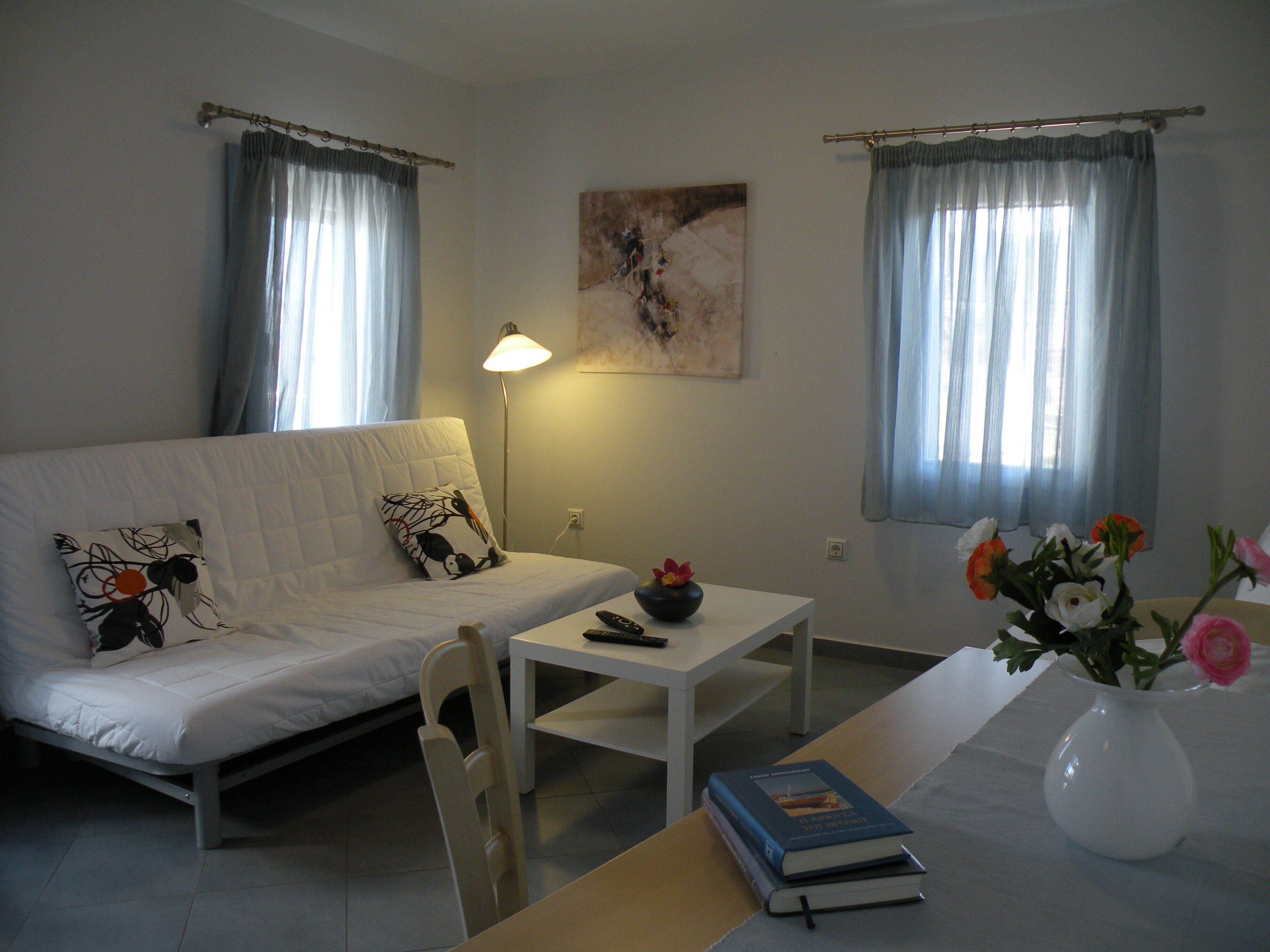 A room with a white sofa, a small table, a lamp, a vase with flowers, and a painting on the wall.