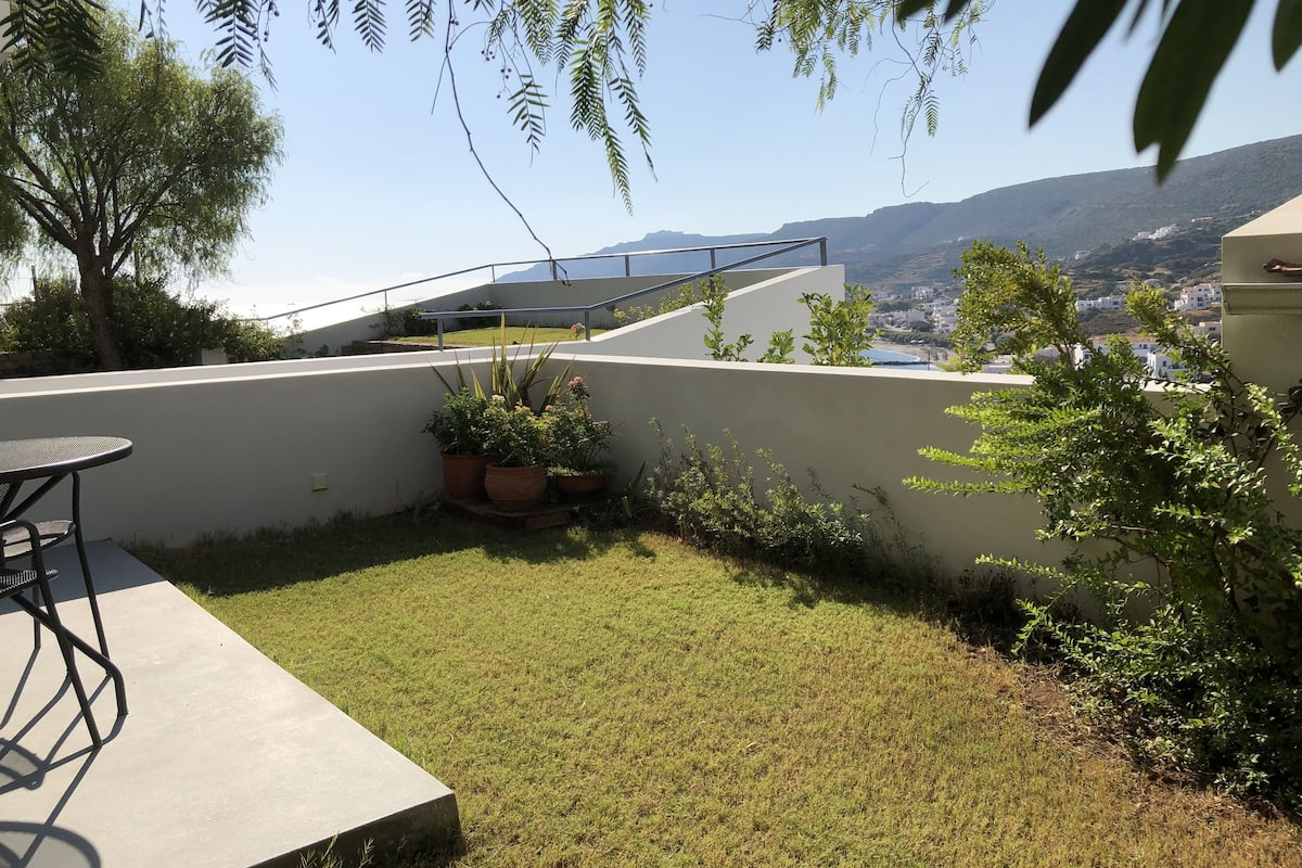 A patio with a concrete table and chairs, surrounded by grass and potted plants. A white wall and a metal railing are visible.