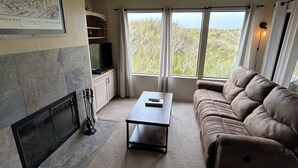 A living room with a fireplace, a sofa, a coffee table, and a large window with a view of the outdoors. - Modern Beach Front Pajaro Dunes 1st Floor Condo - Yards From Beach/Gated Entry (Watsonville)