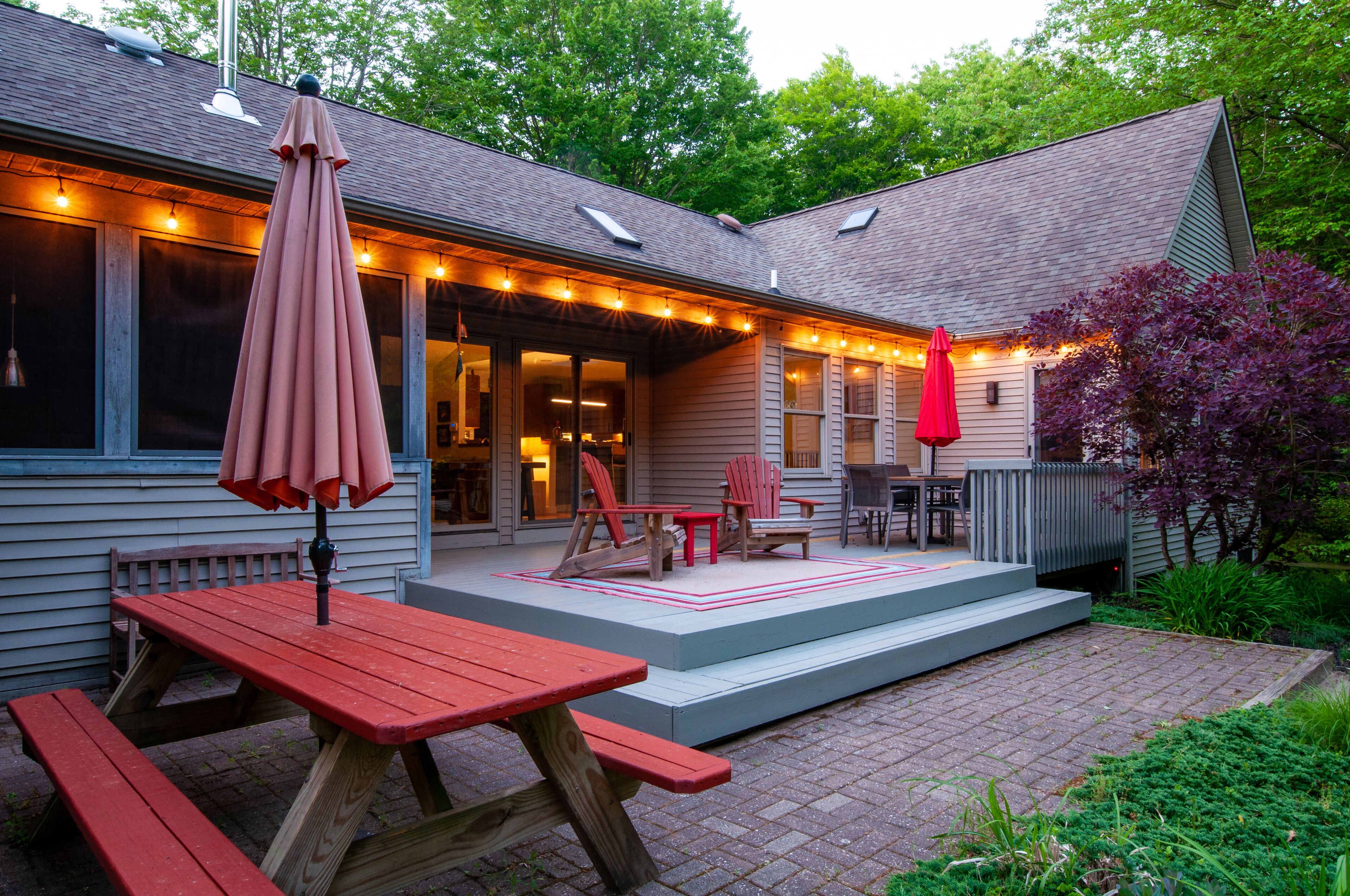 Une terrasse couverte avec des tables et des chaises pour pique-nique, des guirlandes lumineuses et une terrasse en bois qui mène à la maison.