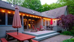 A covered patio with picnic tables and chairs, string lights, and a wooden deck leading to the house. - Wooded Retreat - Steps to the Lake - Close to Town (Fennville)