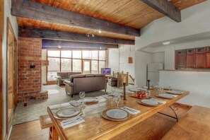 Uma sala de jantar com uma mesa de madeira comprida posta para refeição, uma lareira de tijolos e uma cozinha moderna ao fundo.