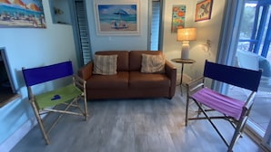A living room with a brown sofa, two folding chairs, a small table with a lamp, and a painting of a beach scene. - Unique Houseboat on Dry Lot-Perfect Romantic Getaway Or Family Friendly & Pet OK (Key Largo)