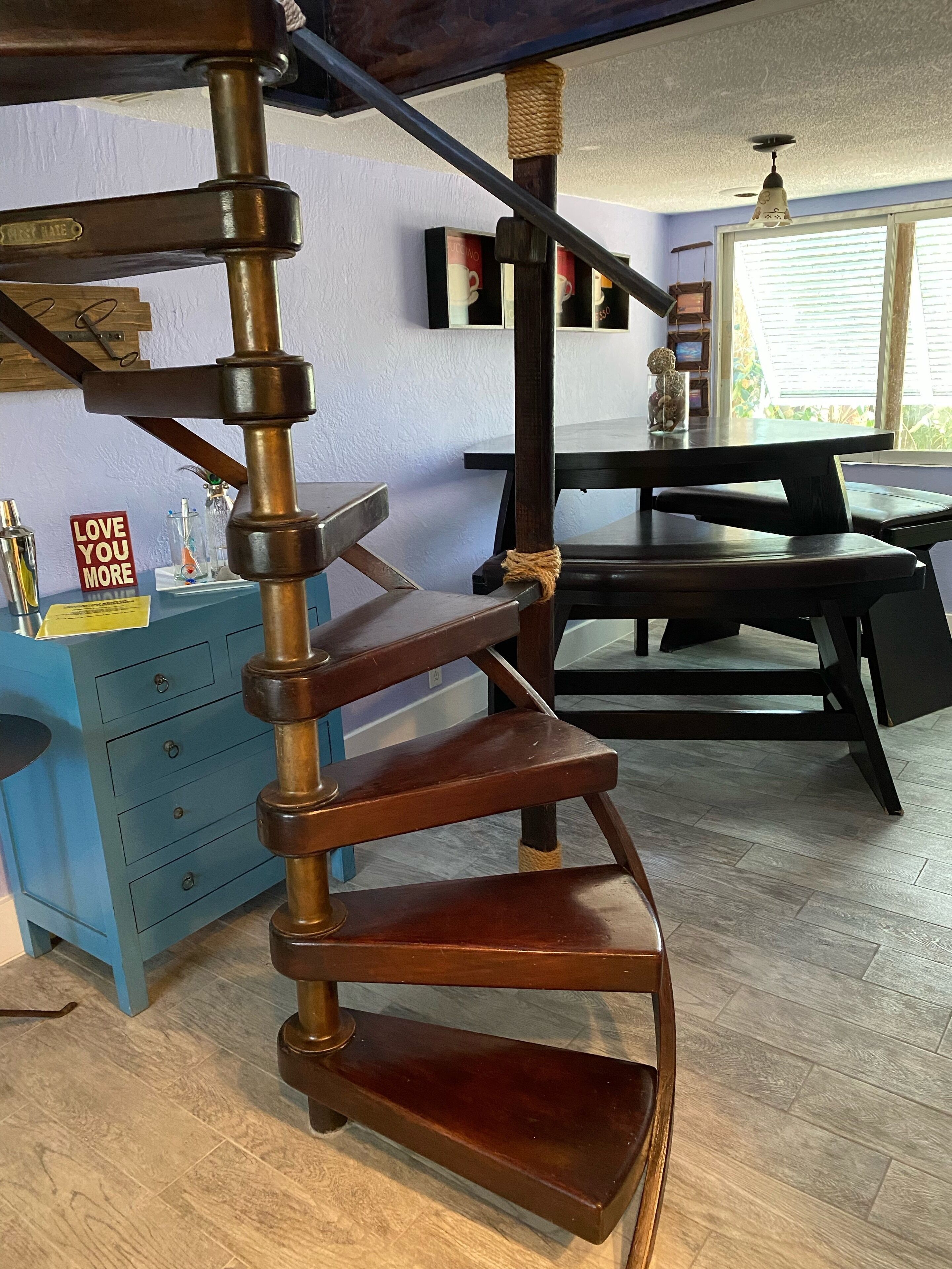 Eine Wendeltreppe mit Holzvertäfelung, eine blaue Kommode mit Schubladen, ein Schild mit der Aufschrift 'LOVE YOU MORE' und im Hintergrund ein Essbereich mit Tisch und Stühlen.