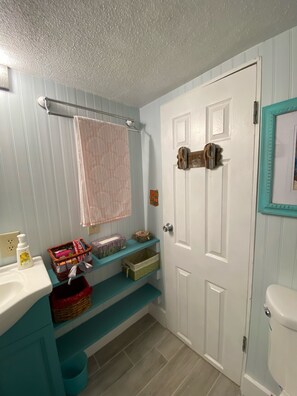 Un baño con puerta blanca, un lavabo con espejo, toallero y estantes con cestas y artículos de aseo.