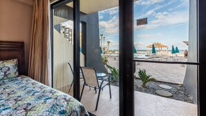 A hotel room with a bed, a chair, and a view of a beach with umbrellas and lounge chairs.
