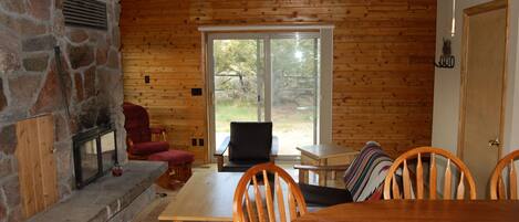 A wooden cabin interior with a stone fireplace, a dining table, chairs, and a large window.