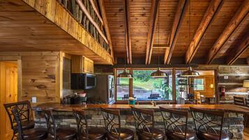 A rustic cabin interior with a stone fireplace, wooden ceiling beams, and a bar area with chairs.