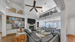 A spacious living room with a large sectional sofa, a fireplace, and a television. There is a balcony with a railing and a view of the ocean.