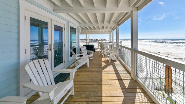 A covered deck with white chairs and tables, overlooking a beach.