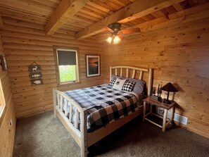 Ein Schlafzimmer einer Holzhütte mit Bett, Nachttisch, Lampe, Deckenventilator und Fenster mit Blick auf Grünflächen.