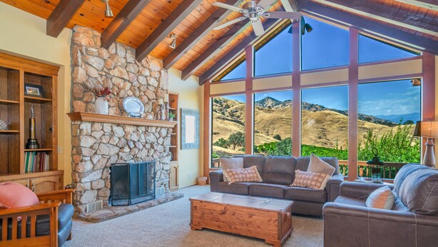 A spacious living room with a stone fireplace, large windows offering a view of rolling hills, and a cozy seating area. - Ranch Getaway in the foothills of the Sierras (Springville)